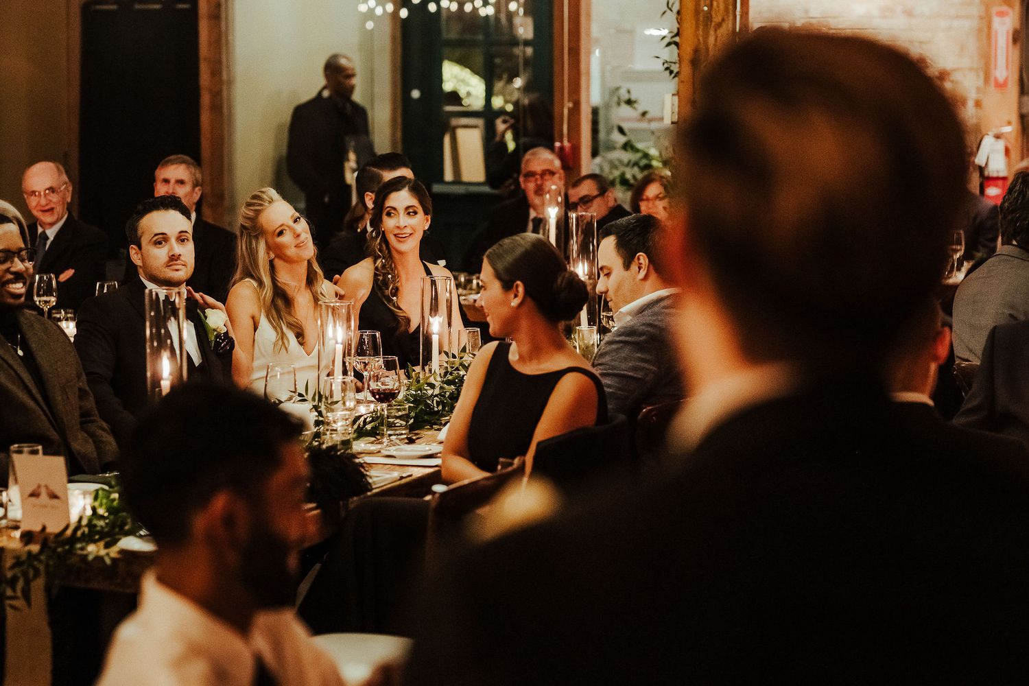 Warm candlelit dinner reception with guests seated at long tables in rustic venue.