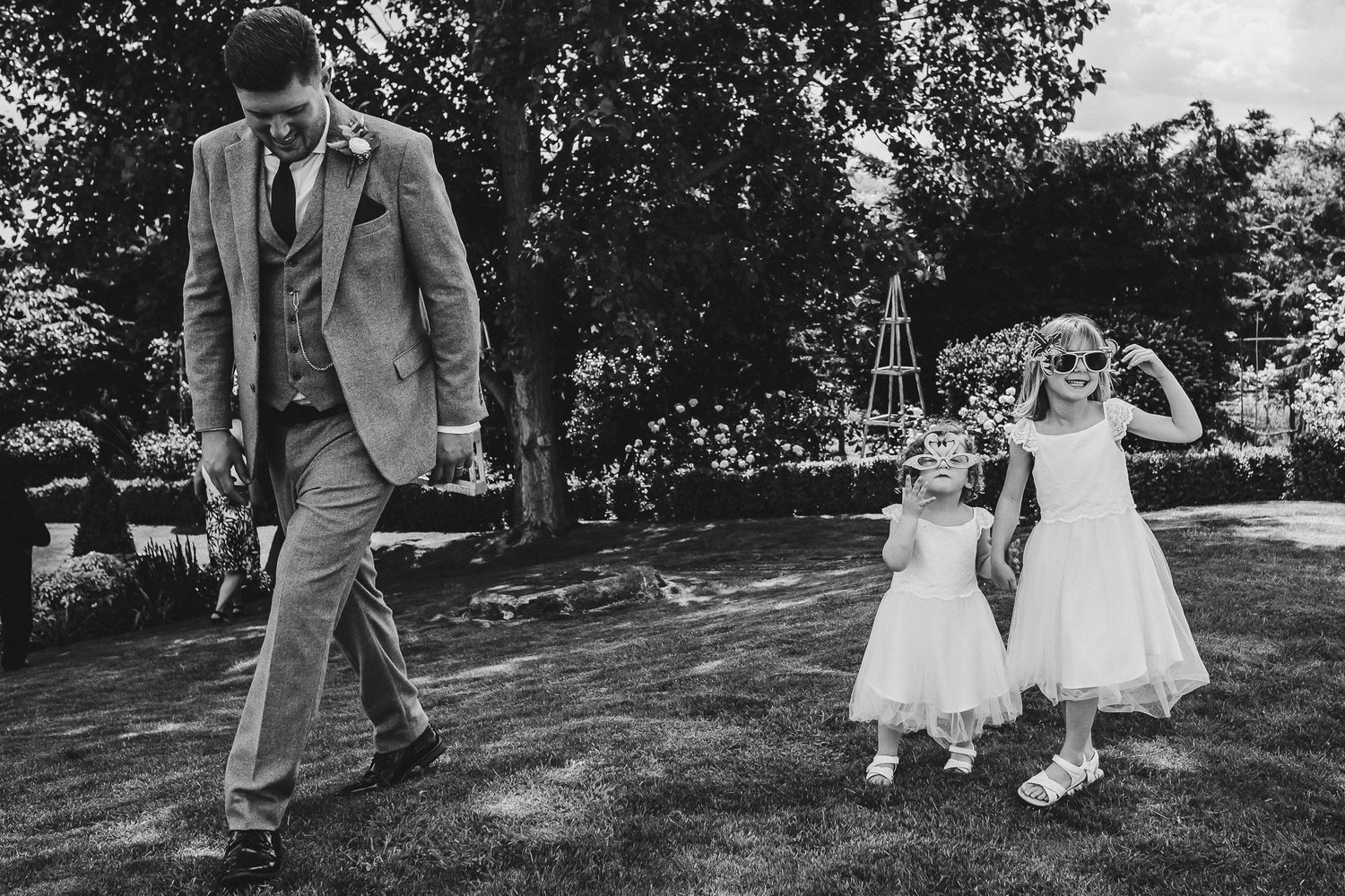 Black and white photo of children in white dresses dancing while adult walks in garden.