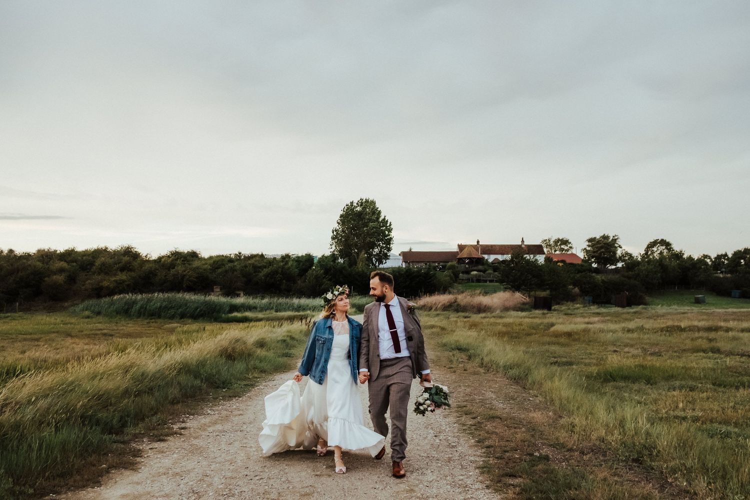 A couple walks down a dirt path through a grassy field on their wedding day at sunset.