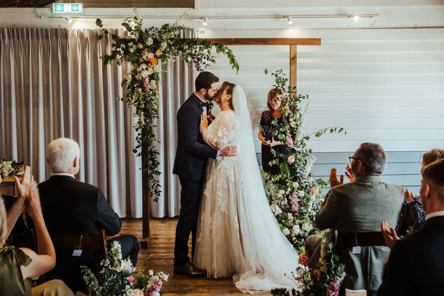Wedding ceremony in a bright indoor venue with floral arch decorations and seated guests.