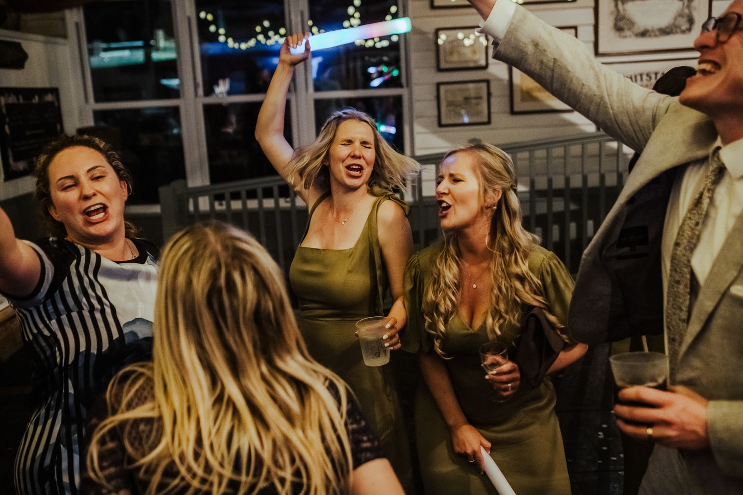 Party guests dance energetically while holding light-up foam sticks against exposed brick walls.