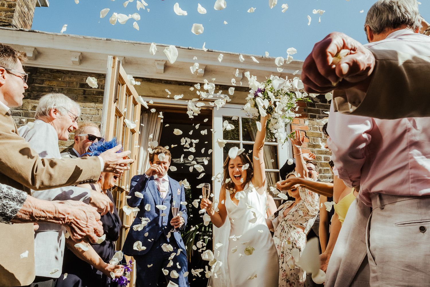 Wedding guests shower confetti on newlyweds outside a rustic venue on a sunny day.