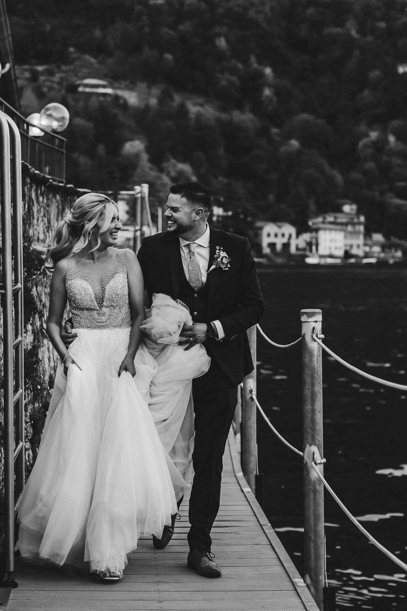 Black and white photo of a couple walking along a waterfront pier in wedding attire.