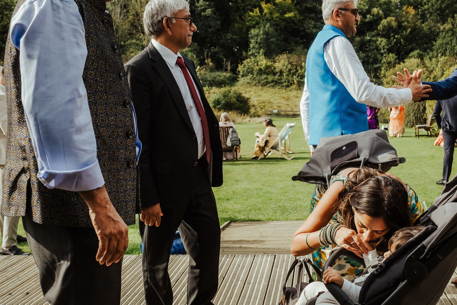 People gathering in a park on a wooden deck while someone bends down to interact with a child in a stroller.