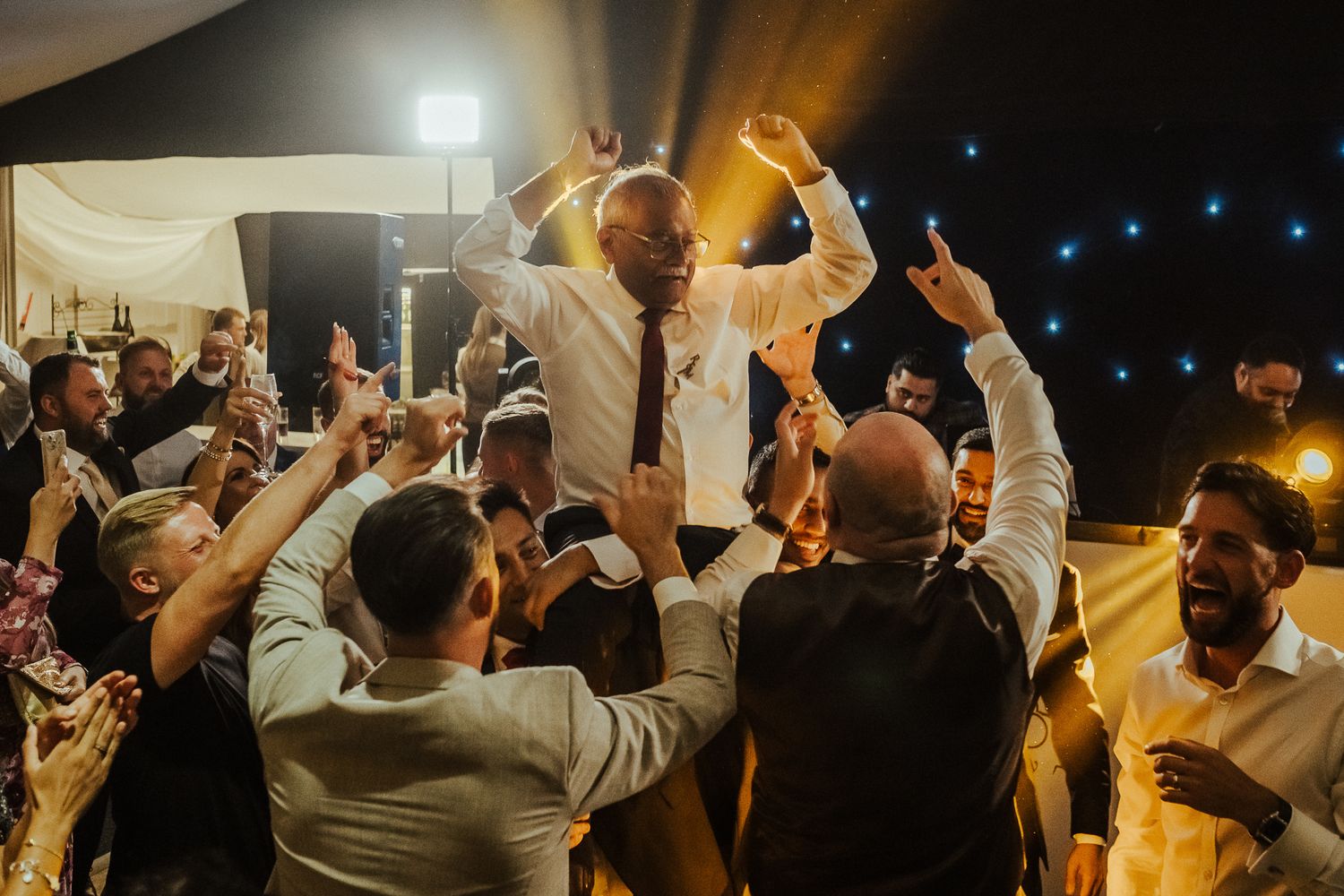 Wedding guests lift and celebrate with enthusiastic guest in dress clothes at nighttime reception party under tent.
