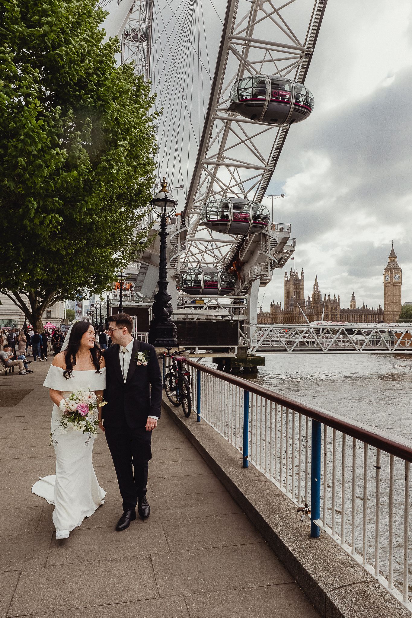 Two people celebrate with London Eye and Houses of Parliament visible along River Thames on cloudy day.