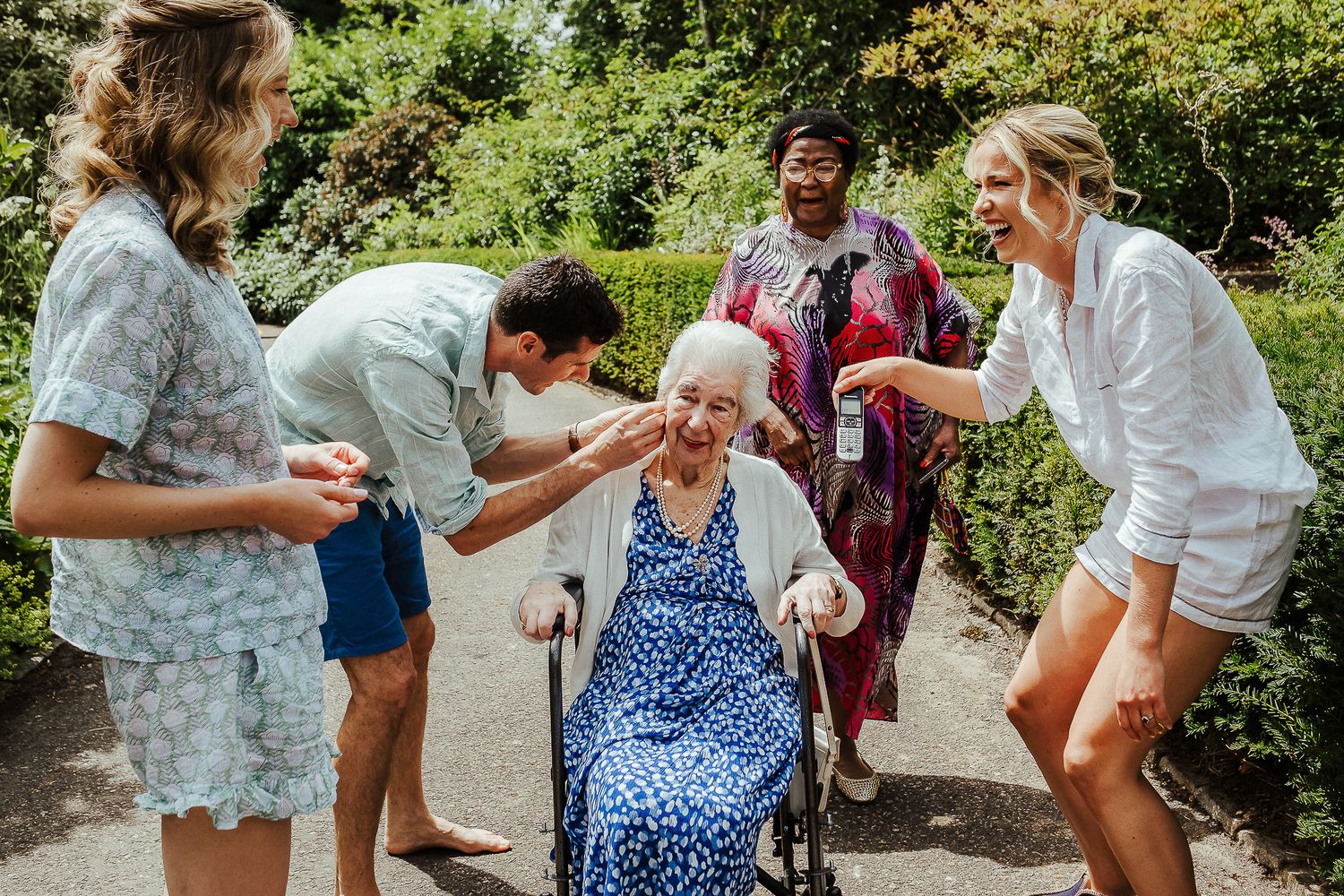 Group gathers around elderly woman in blue dress outdoors during summer celebration.