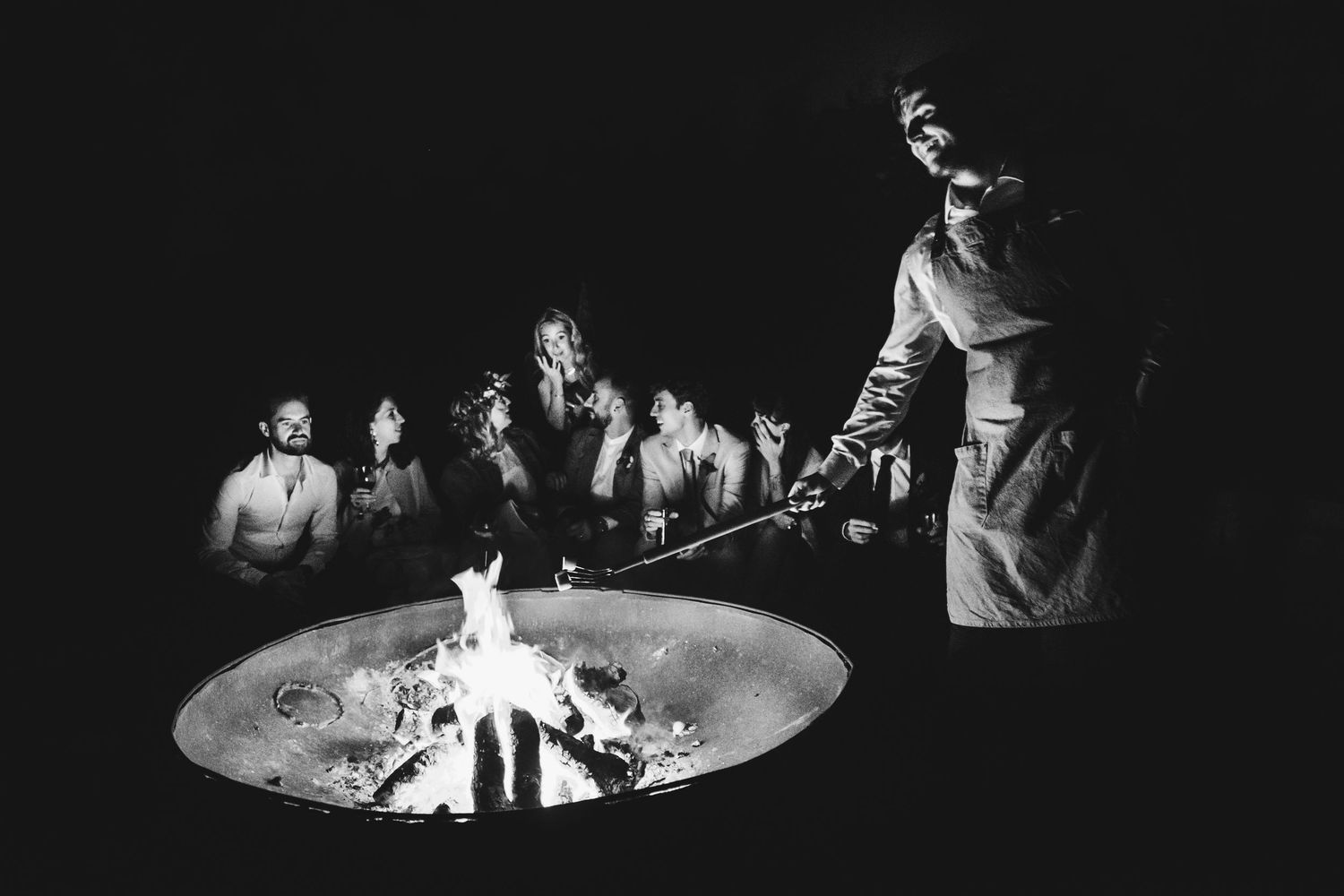 People gather around a fire pit at night creating a dramatic black and white scene.
