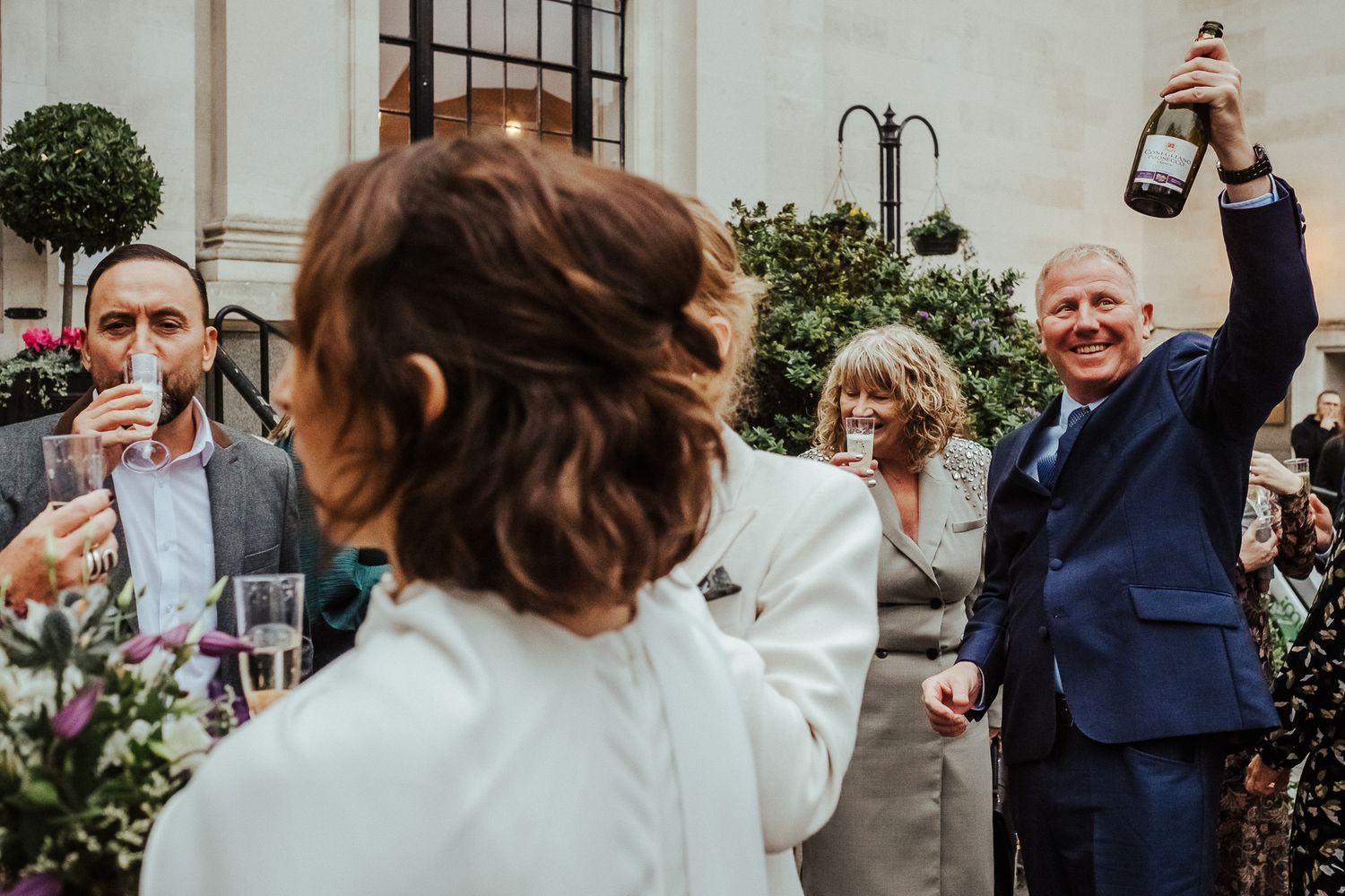 Wedding guests raise champagne bottles in celebration outdoors.