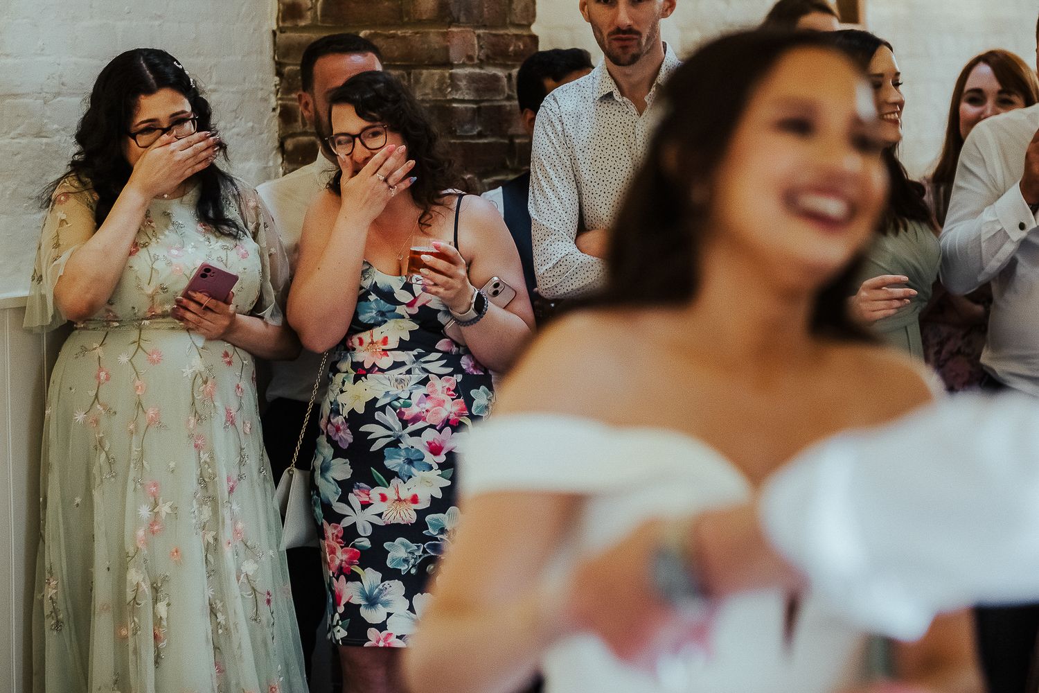 Guests at a wedding reception reacting with joy and laughter against a brick wall backdrop.