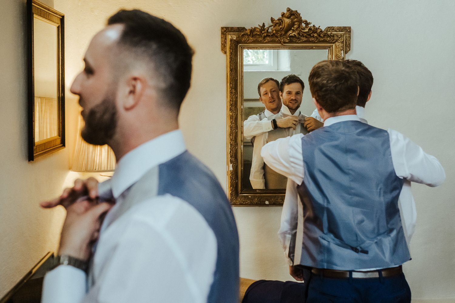 Groomsmen adjust their ties while getting ready in front of an ornate gold-framed mirror.
