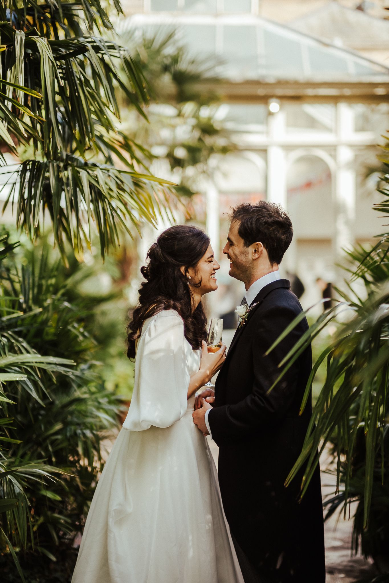 Romantic wedding couple embracing in greenhouse surrounded by lush tropical plants.