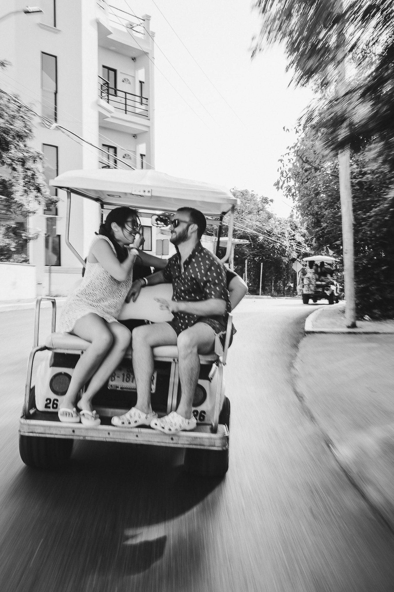Black and white photo of couple riding in golf cart on sunny street.