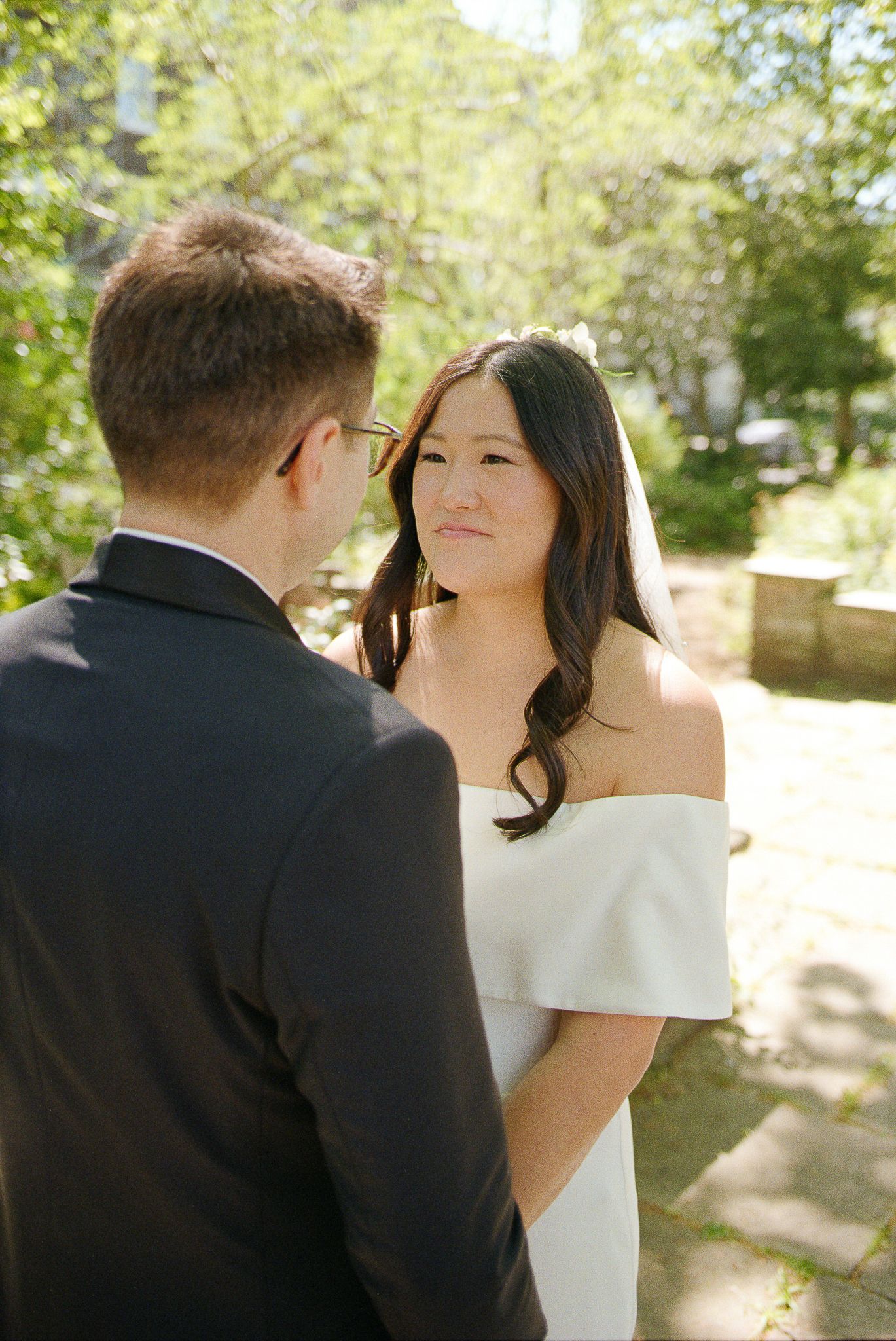 A bride in an elegant off-shoulder white gown gazes lovingly at her groom during an outdoor wedding ceremony.