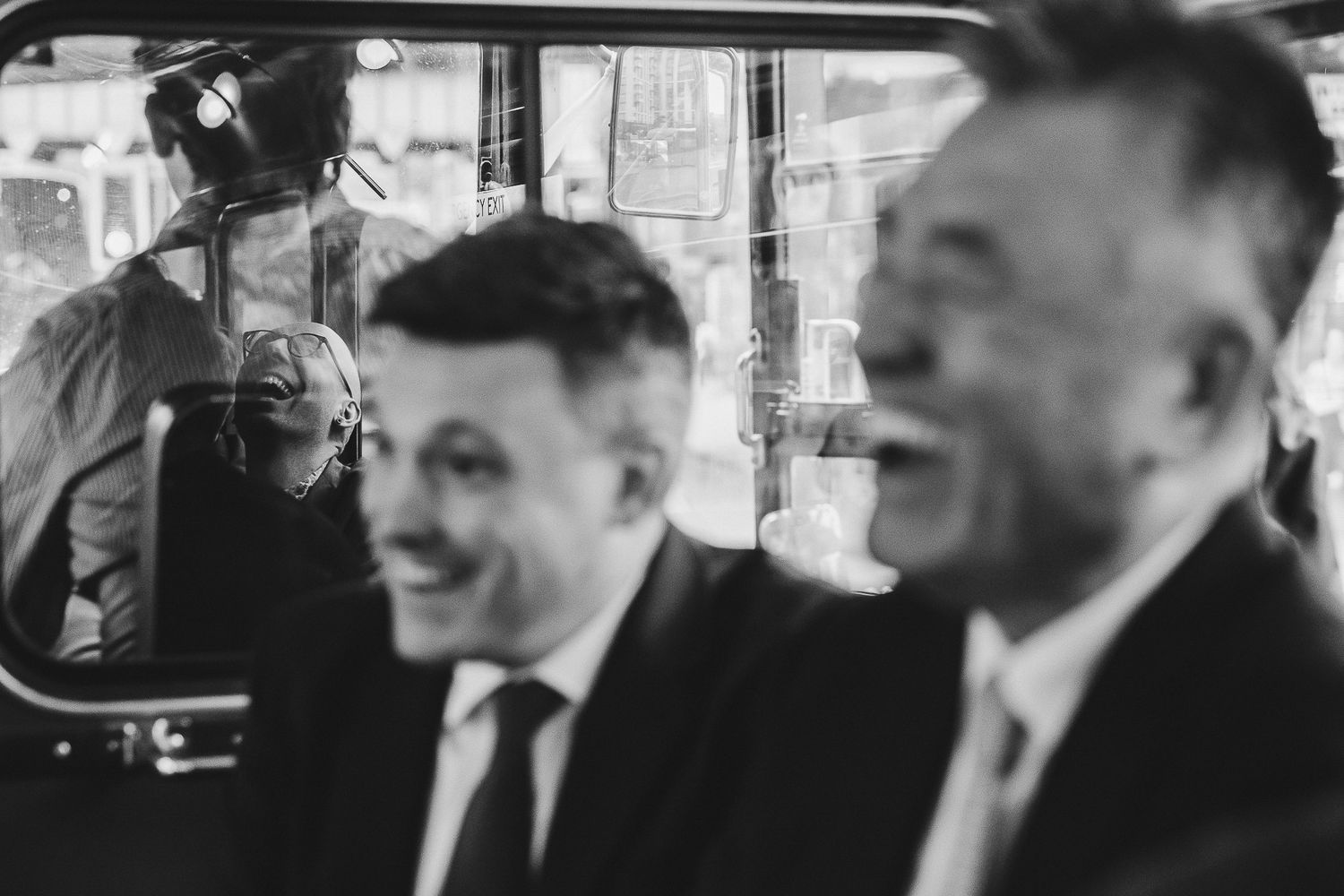 Black and white photograph taken inside a car with two men in suits sitting in the rear seats smiling.