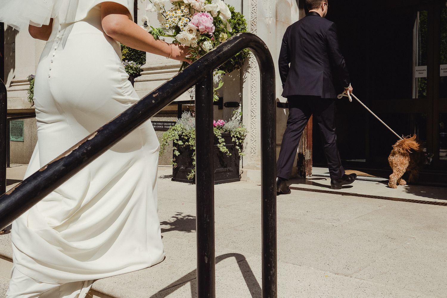 Person in white dress ascending stairs with flower bouquet while someone walks dog in background on city street.