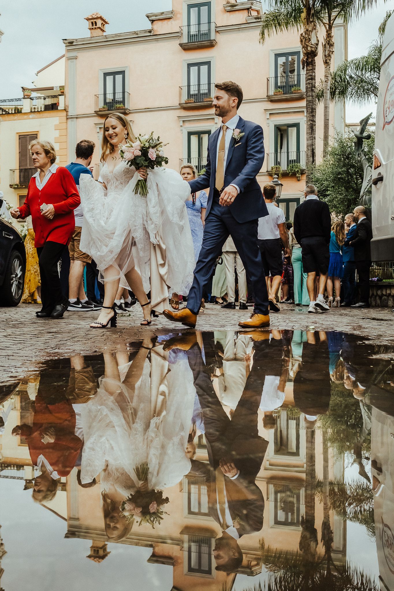 Wedding couple walks past elegant building with their reflection mirrored in a puddle on cobblestone street.
