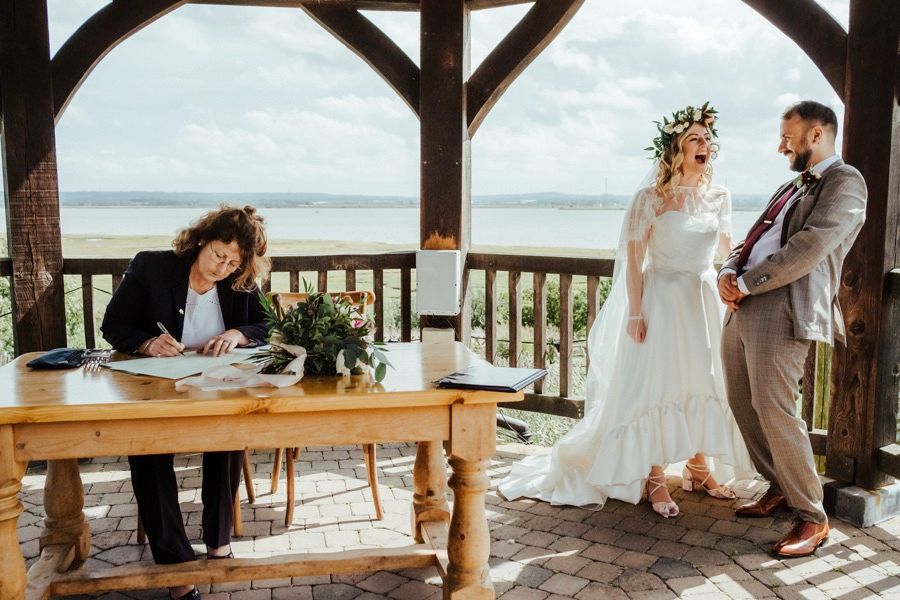 Newlyweds stand on a rustic wooden deck overlooking a lake while signing their marriage certificate.