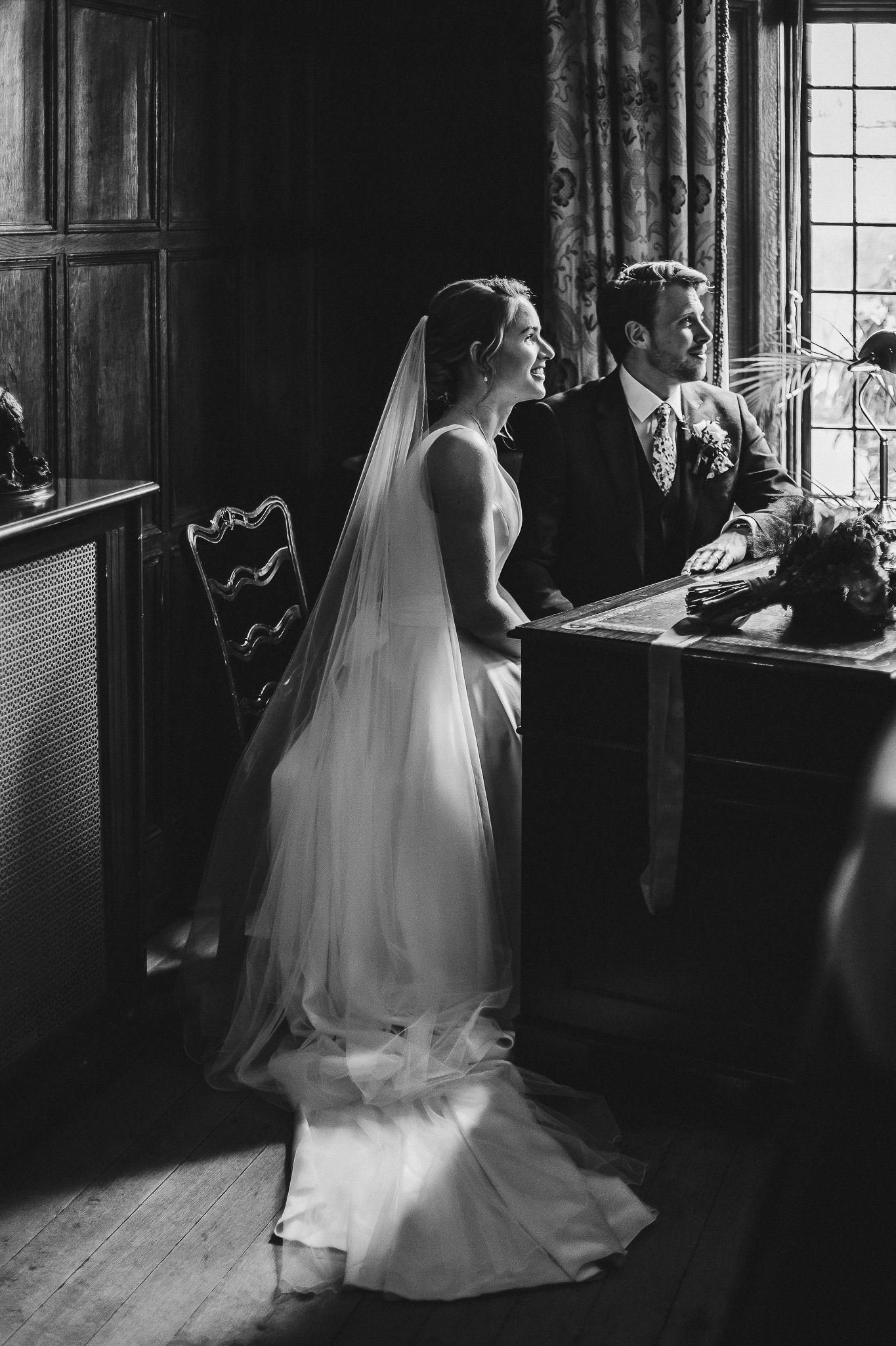 Black and white portrait of newlyweds by window in elegant vintage room.