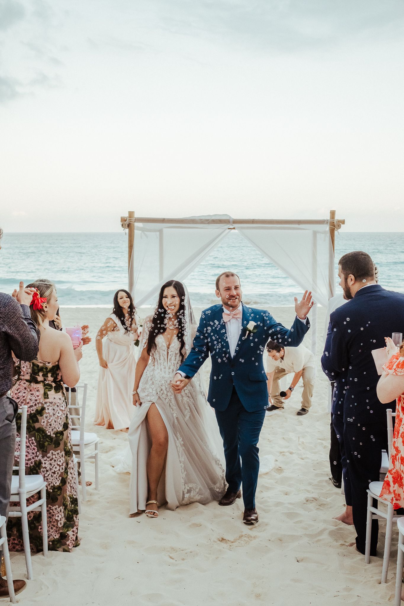 Newlyweds walking down beach aisle while guests celebrate with confetti.