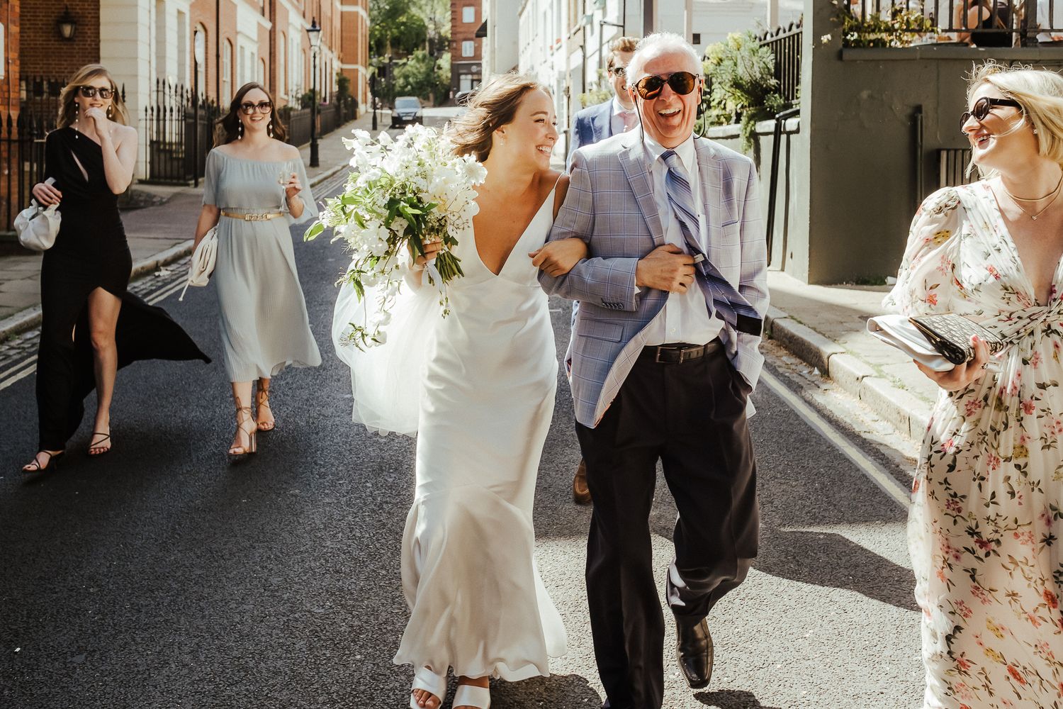 A group walks down a sunny street during a wedding celebration carrying bouquets.