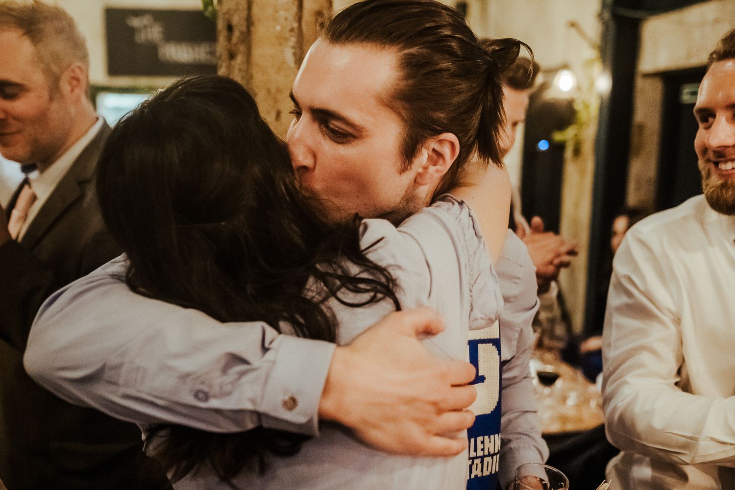 Friends embrace warmly during celebration while holding drinks at indoor venue.