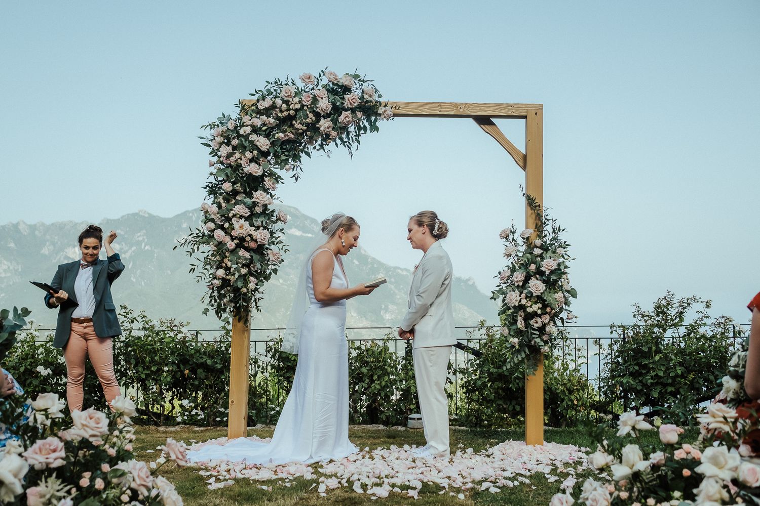 Wedding ceremony under wooden arch decorated with pink and white roses against coastal backdrop.