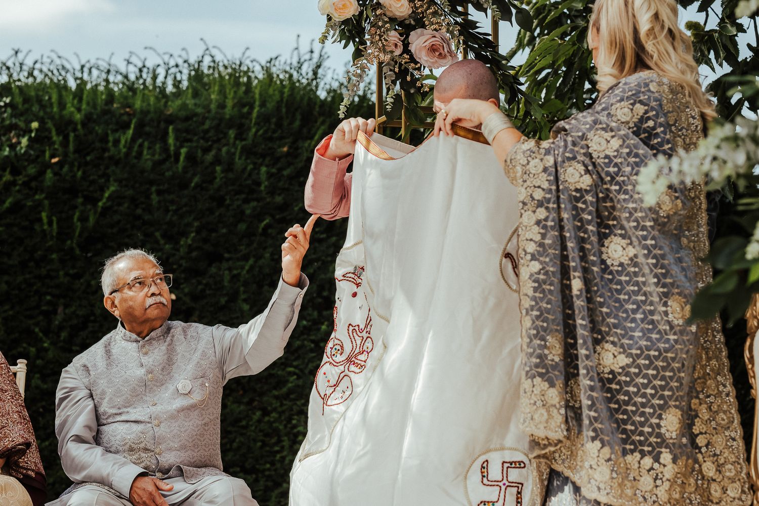 A joyful outdoor gathering with elegant formal attire and floral decor beside a lush green hedge.