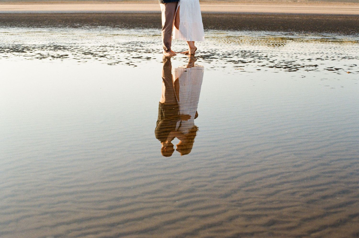 Legs and reflection of someone in white dress standing at the edge of calm water during golden sunset.