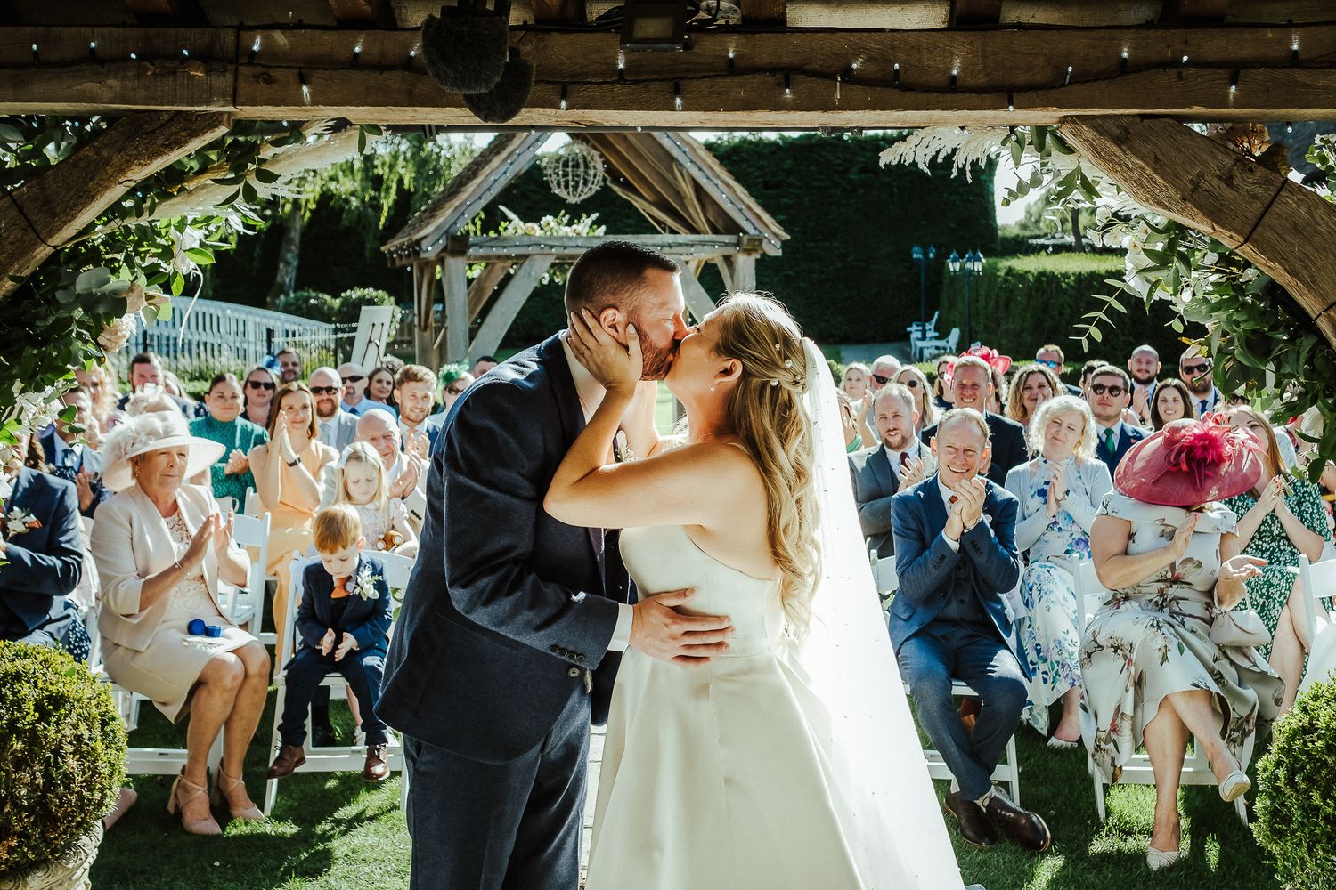 Newlyweds share first kiss under gazebo surrounded by cheering guests.