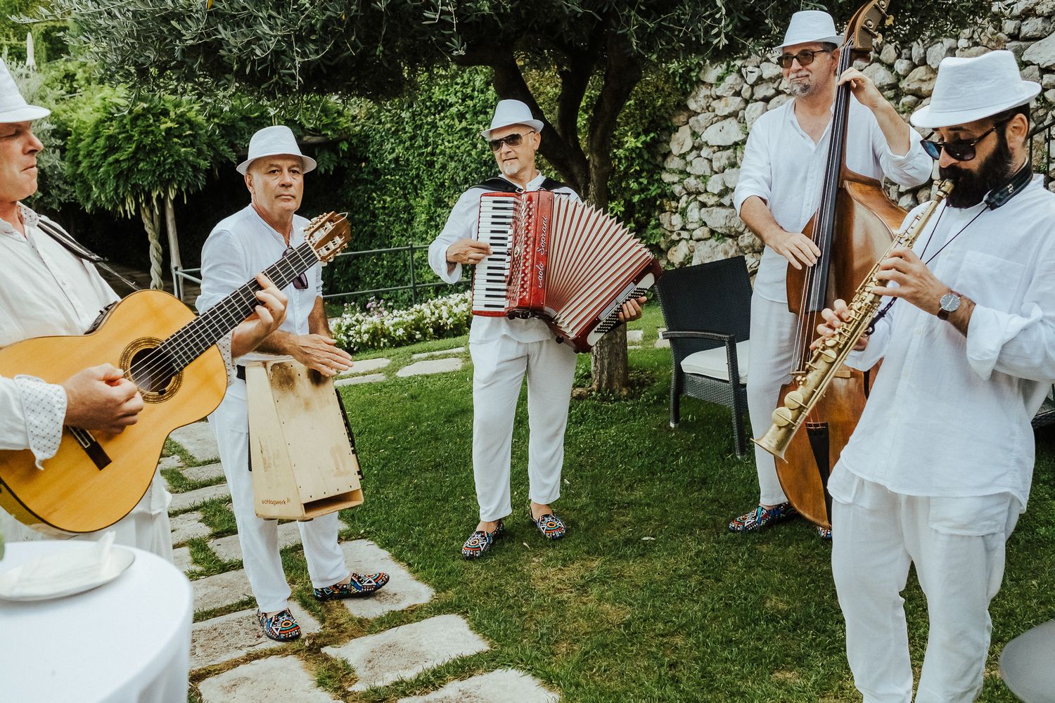 Musicians in white outfits performing with various instruments during an outdoor garden event.