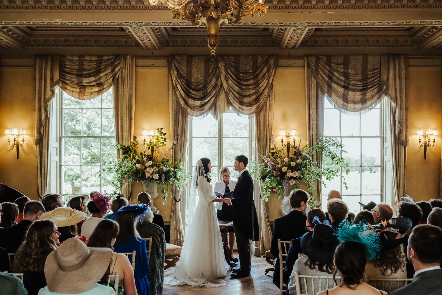 Elegant wedding ceremony in a grand room with ornate windows, chandeliers and floral arrangements.