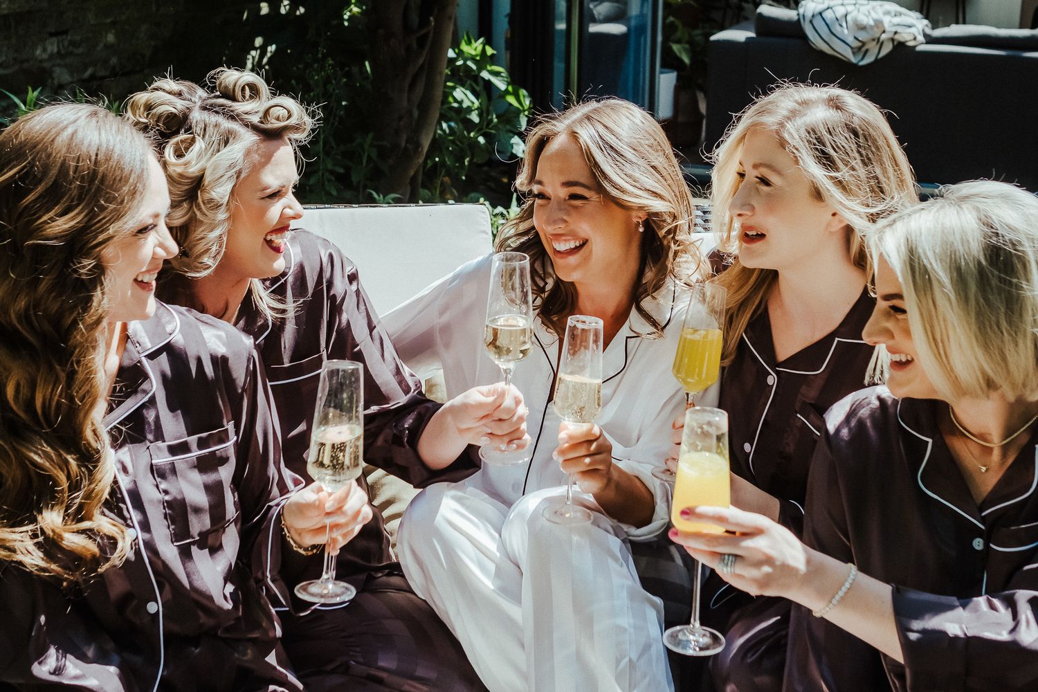 Wedding party members laugh together while holding champagne glasses outdoors.