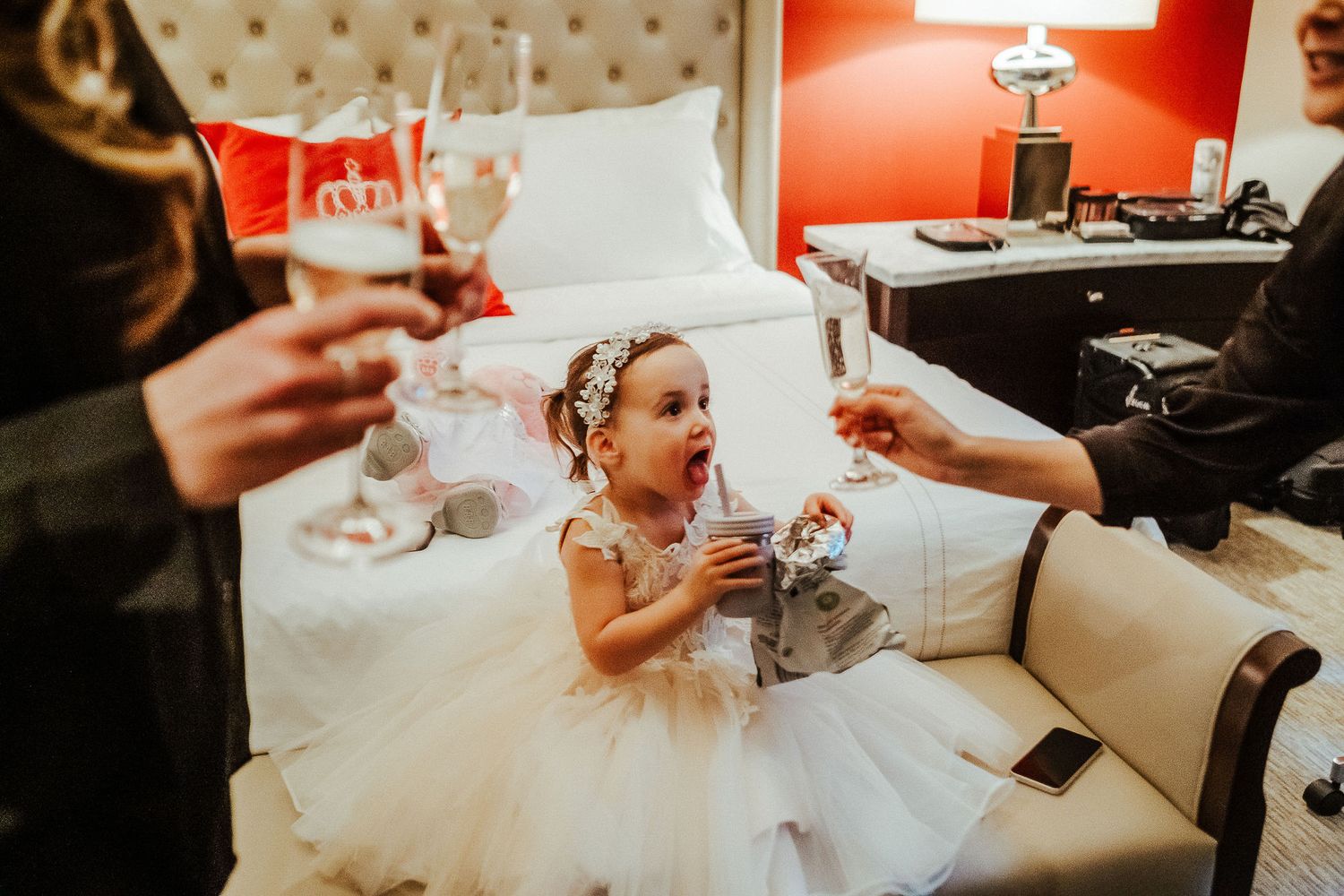 A flower girl in a white tulle dress sits on a bed during wedding preparations.