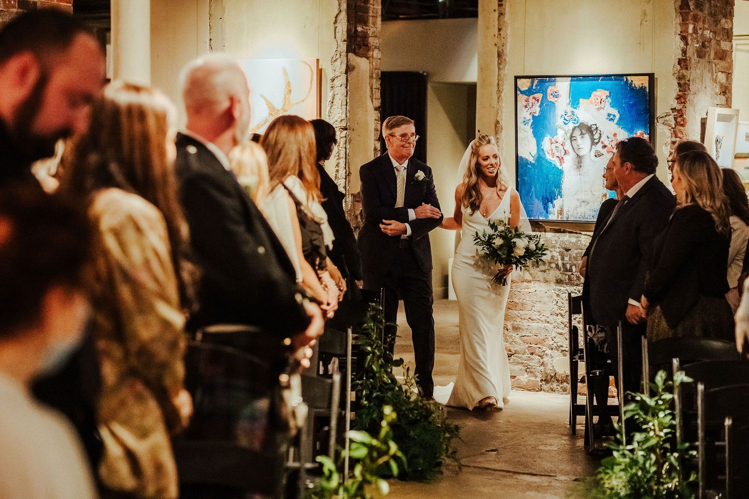 Wedding ceremony in rustic venue with guests lined up along aisle decorated with greenery.
