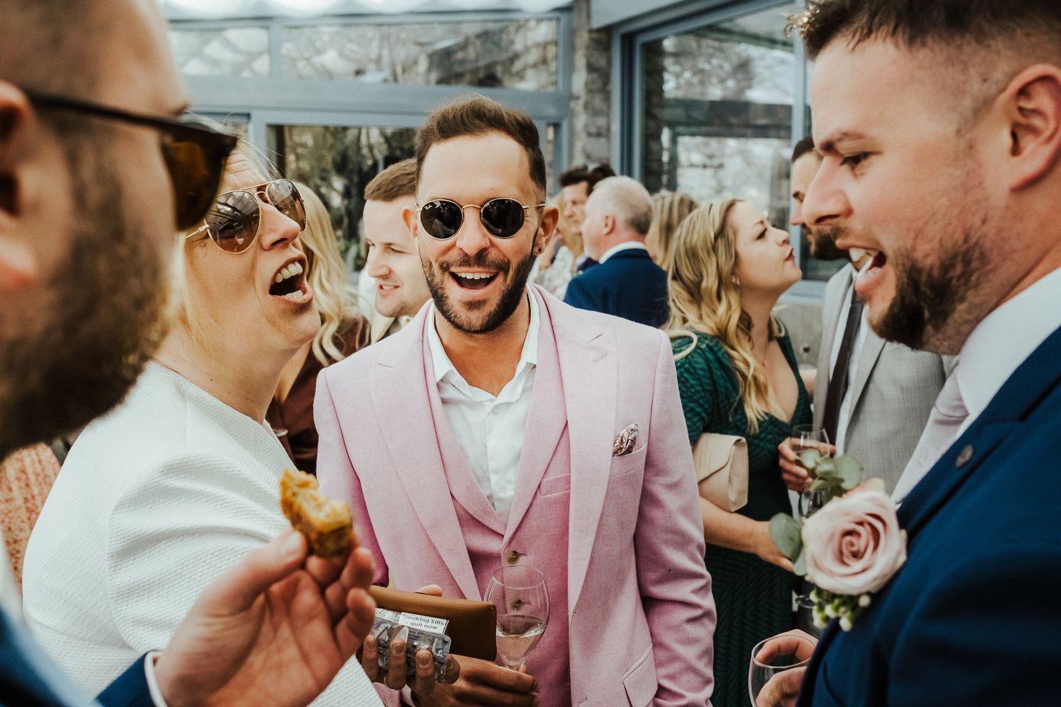 Guests in stylish attire share drinks and laughter during an outdoor wedding reception.