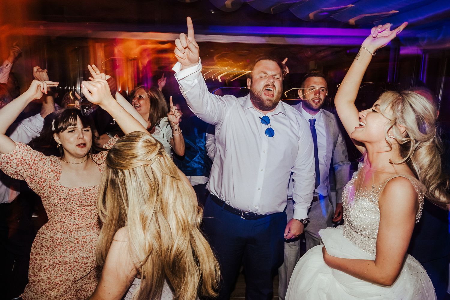 Wedding attendees dancing with raised arms under vibrant reception lighting.
