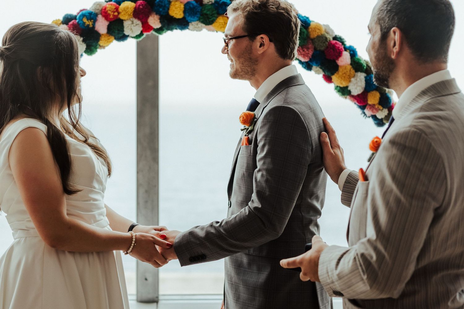 Couple holds hands during wedding ceremony under colorful balloon arch with ocean view.