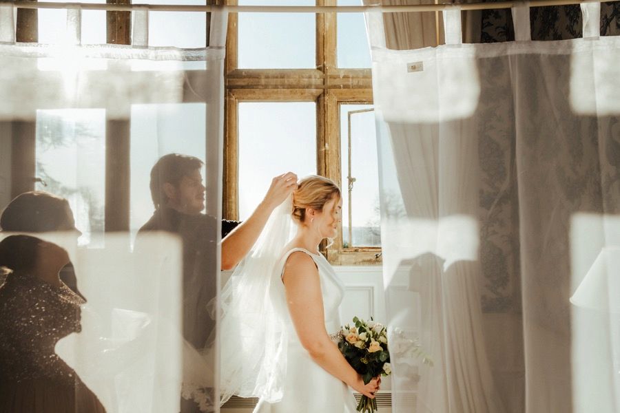 Bride in lace dress getting ready by window with sunlight streaming through sheer curtains.
