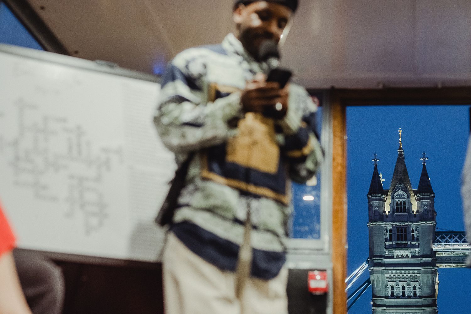 A blurry figure stands in front of a whiteboard with Tower Bridge visible through a window in the background.