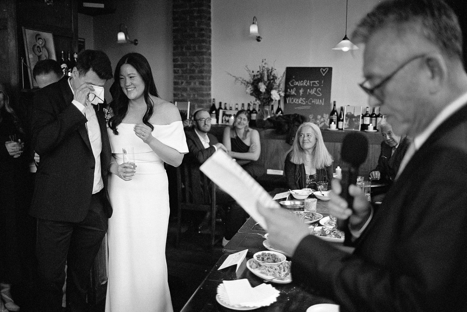 Black and white photo of wedding reception guests mingling and dining in an intimate restaurant venue with exposed brick walls.
