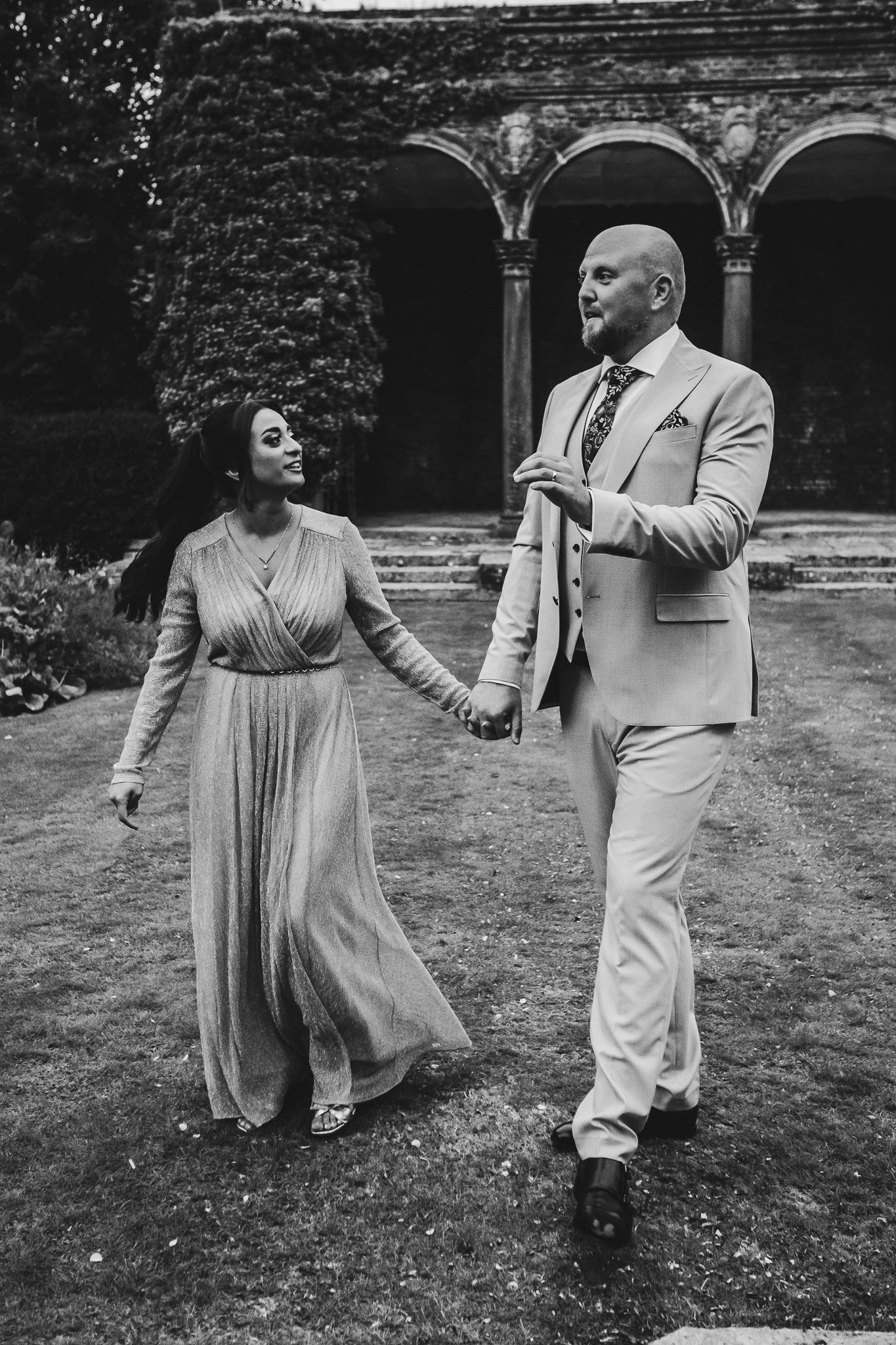 A couple dancing together in formal attire on a lawn with an arched stone building visible in the background.