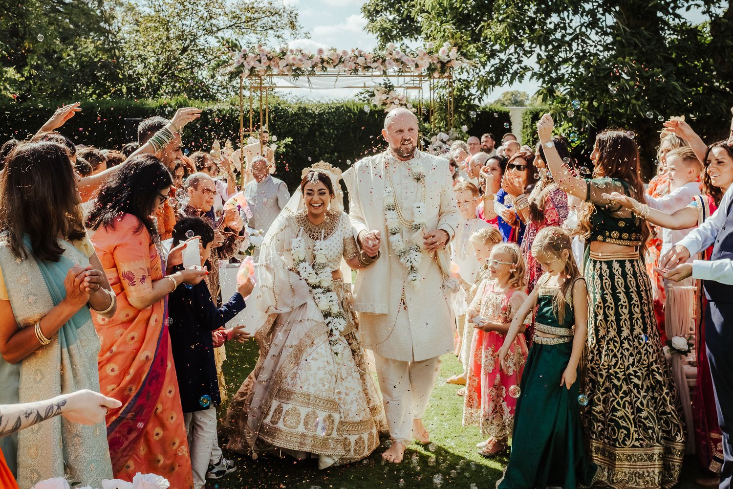 Guests throw flowers and celebrate during a colorful multicultural outdoor wedding ceremony under a floral arch.