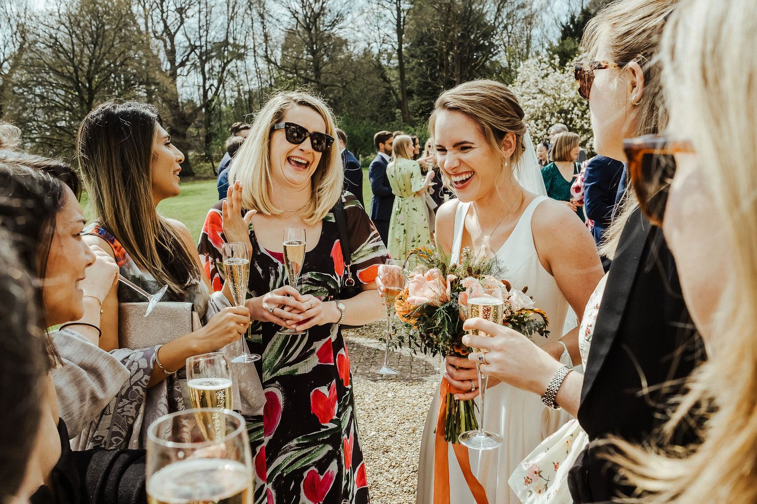 Group of wedding guests sharing laughter while holding champagne glasses in sunlit garden.