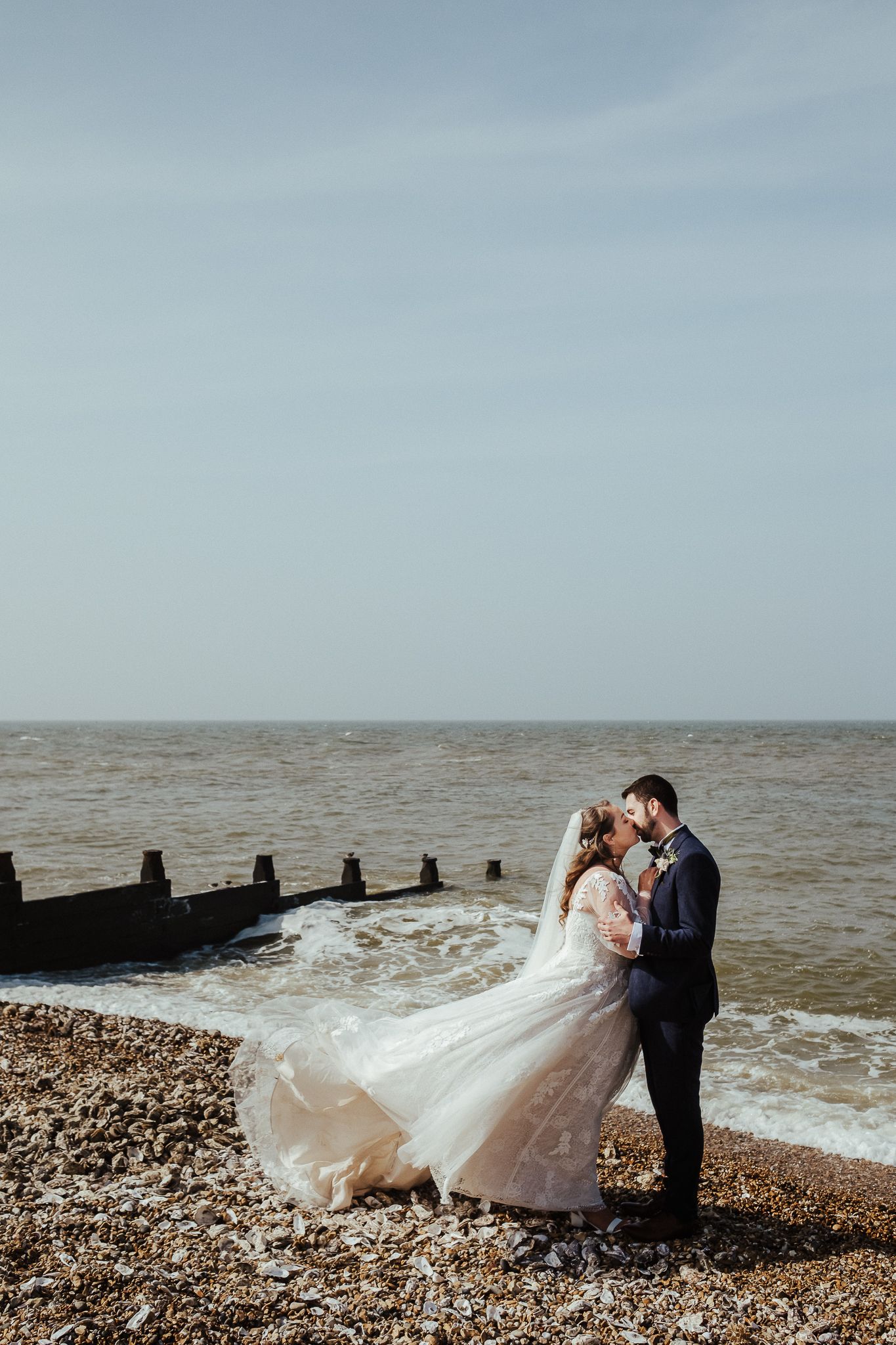 Newlyweds share an intimate moment by wooden groynes on a pebble beach.