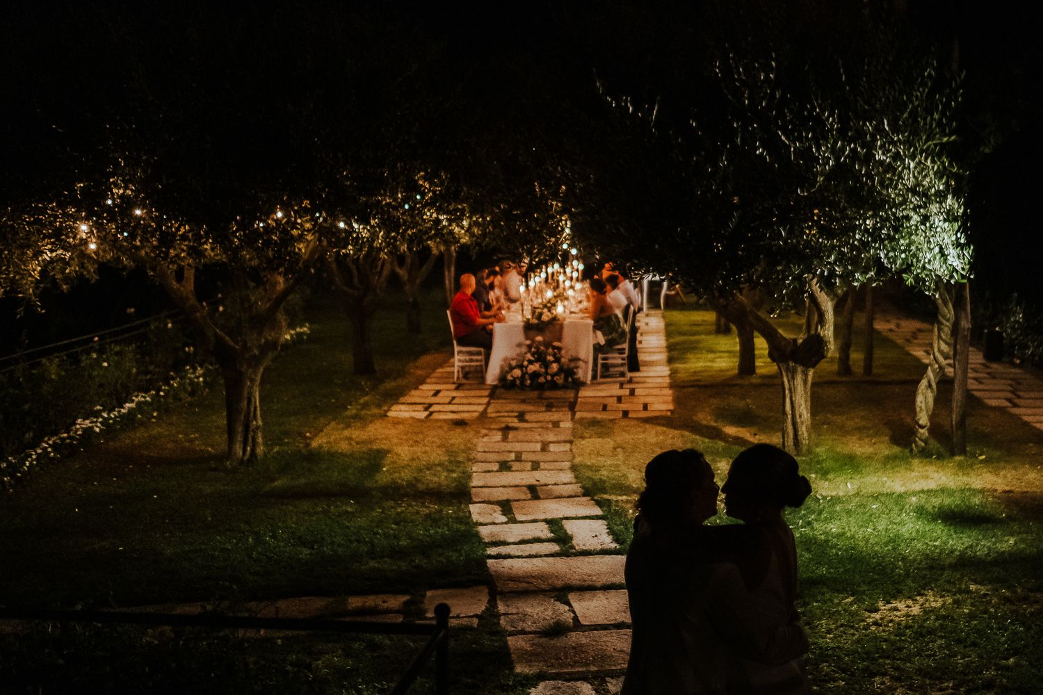 Silhouetted couple watches distant dinner party under illuminated trees.