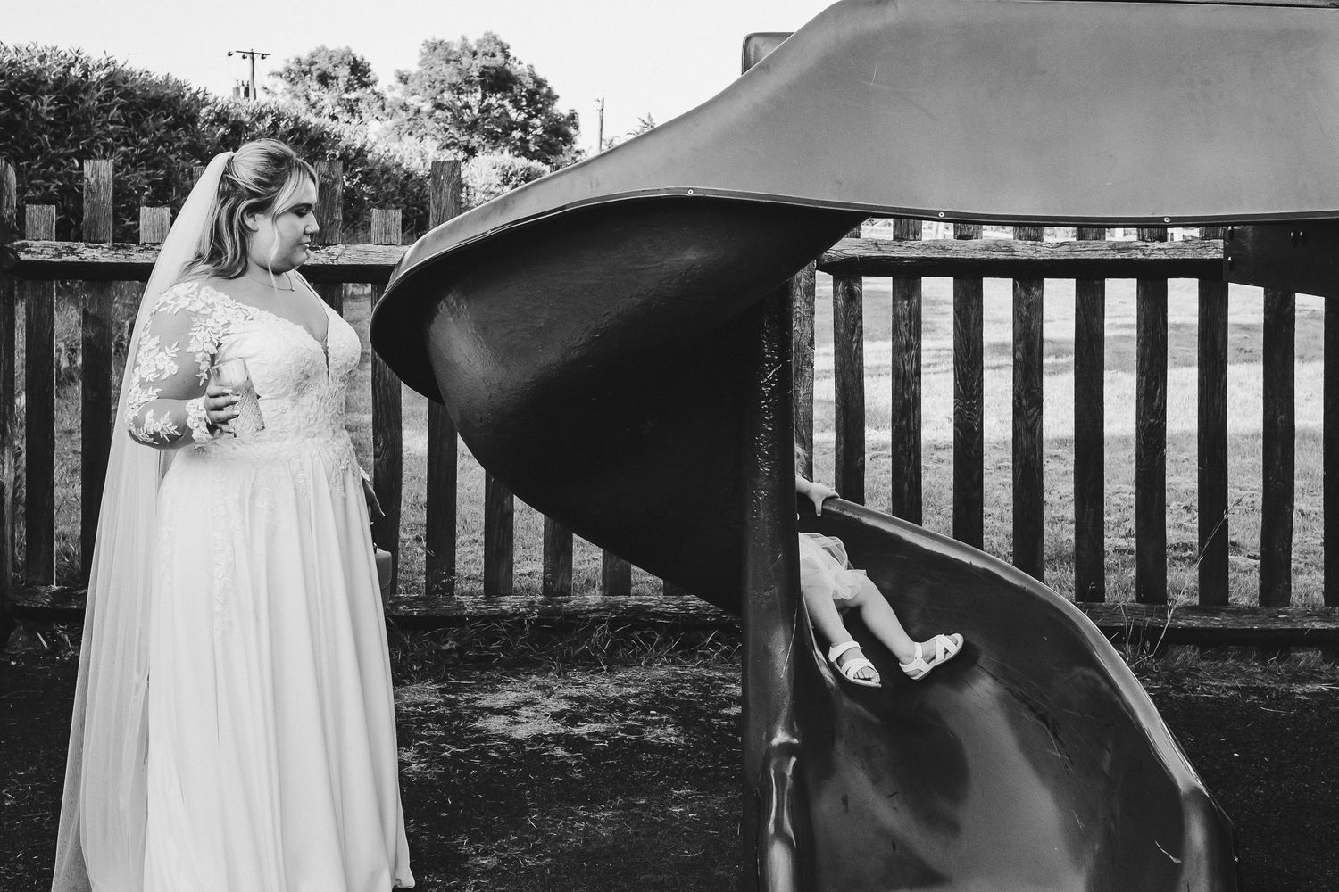 A bride in a white wedding dress peers into a curved black playground slide at an outdoor venue.