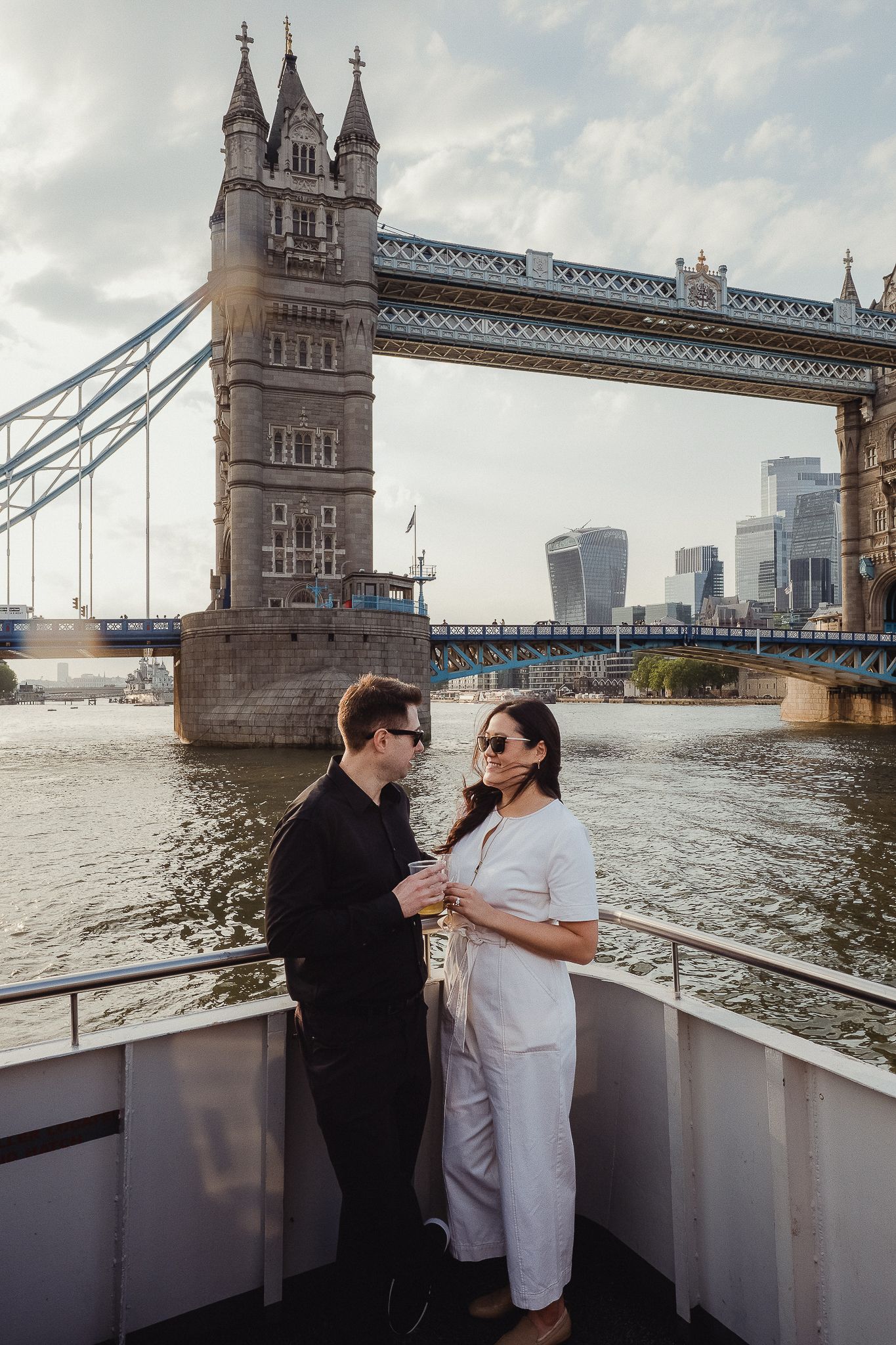 A couple stands on a boat deck with London's iconic Tower Bridge and modern city skyline in the background.