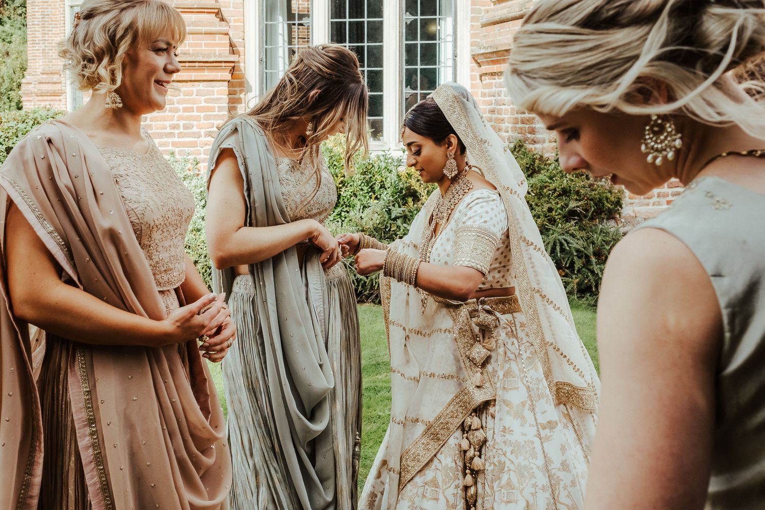 People in soft neutral-toned dresses gather outdoors for a wedding ceremony near a brick building.