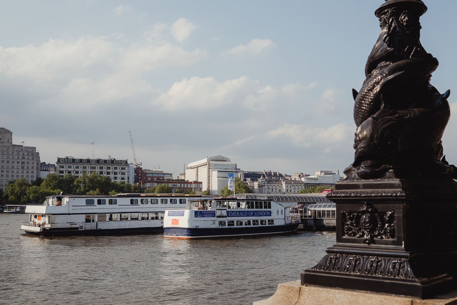 A tourist boat cruises on the Thames River in London with ornate bridge details and city buildings in the background.